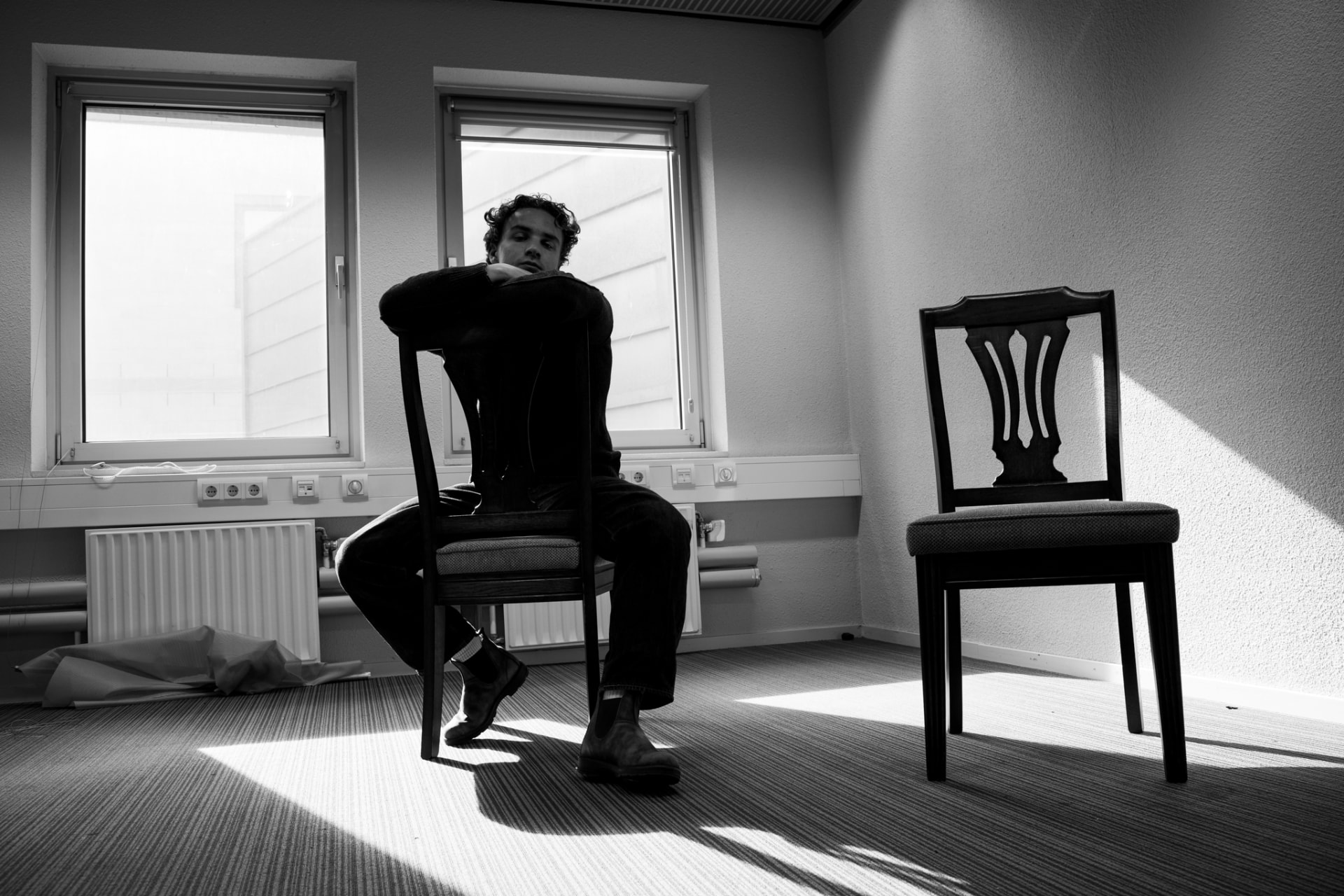 Zwart-witfoto: een figuur leunt voorover op een stoel in een lichte, lege ruimte, naast een lege stoel.